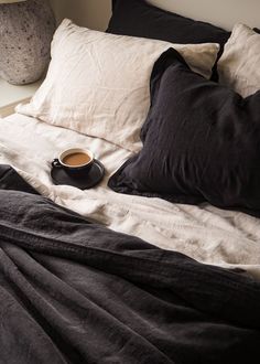 a cup of coffee sits on the edge of an unmade bed with black linens