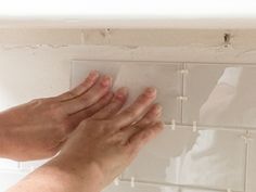 two hands reaching for something on a white tile wall above a sink in a bathroom