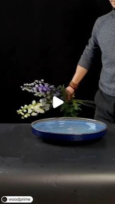 a man arranging flowers in a blue bowl on a black table with a video screen