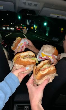 two people holding sandwiches in their hands while sitting in the back seat of a car