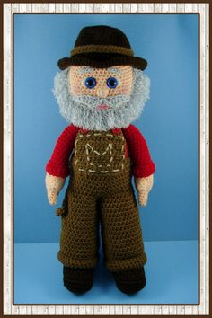 a crocheted doll wearing overalls and a hat with a beard, standing against a blue background