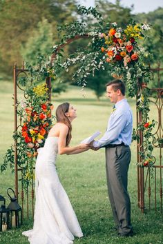 a bride and groom hold hands under an arch decorated with orange, yellow and red flowers