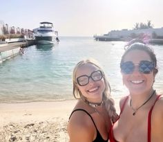 two women standing next to each other on a beach with boats in the water behind them