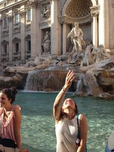 two women standing in front of a fountain with their hands up to catch the frisbee