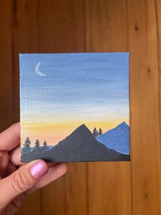 a hand holding up a small piece of art with mountains in the background at sunset