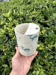 a hand holding up a cup in front of some bushes and trees with blue whale designs on it