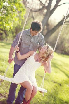 a man and woman sitting on a swing in the grass with their hands behind them