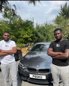 two men standing next to each other in front of a black car with their arms crossed