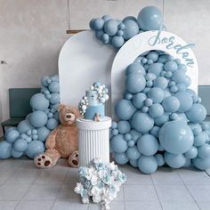 a teddy bear sitting in front of a balloon arch with blue balloons on the wall