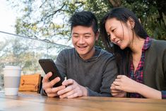 a man and woman sitting at a table looking at a cell phone with a cup of coffee in front of them