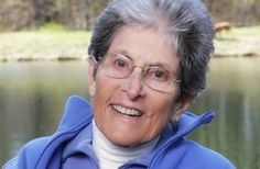 an older woman wearing glasses and a blue jacket smiles at the camera while standing in front of a body of water