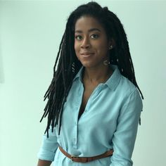 a woman with dreadlocks standing in front of a white wall wearing a blue shirt