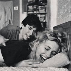 two people are laughing while laying on a bed with their arms around each other and one person is looking at the camera