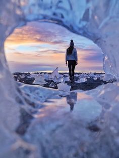 a woman standing in an ice cave looking out at the ocean and sunset behind her