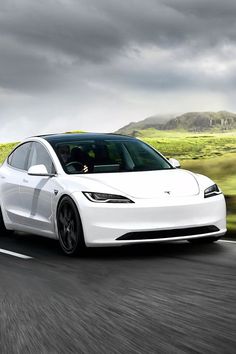 a white tesla car driving down the road in front of some hills and grass on a cloudy day