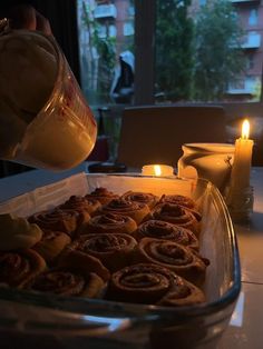 a person pours coffee into a pan filled with cinnamon rolls on a table next to candles