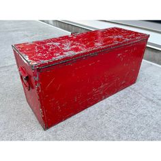 an old red metal box sitting on the ground next to a window sill,