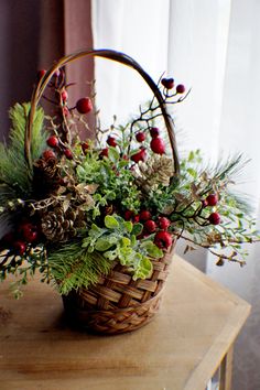 a basket filled with greenery and pine cones on top of a wooden table next to a window