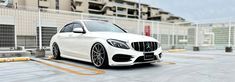 a white mercedes benz is parked in front of a fenced parking lot with buildings behind it