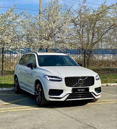 a white volvo suv parked in a parking lot next to a fence and trees with blossoming branches