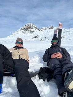 two snowboarders sitting in the snow with their feet propped up and looking at their cell phones