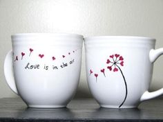 two white coffee cups with red hearts painted on them, one has the words love is in the air