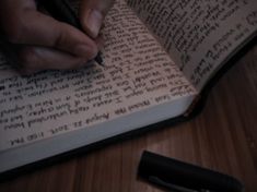 a person writing on an open book with a pen in their left hand and a black object next to it