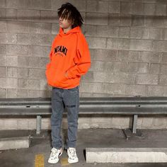 a young man in an orange hoodie stands against a wall with his hands on his hips