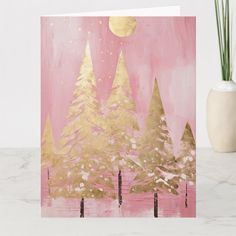 a pink and gold christmas card with trees in the foreground on a marble surface