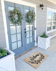 two wreaths on the front door of a house with welcome mat and potted plants