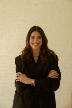 a woman with her arms crossed standing in front of a white wall wearing a black suit and gold watch