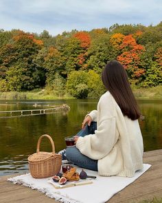 a woman sitting on the edge of a lake next to a picnic basket and food