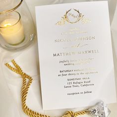 a white and gold wedding card on a table next to a glass with a candle