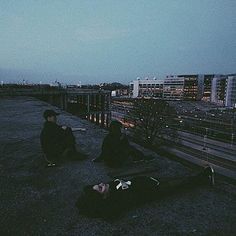 two people sitting on the ground in front of a train track at night with buildings in the background