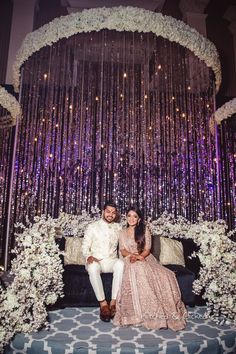 two people sitting on a couch in front of a purple and white backdrop with flowers