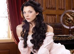 a woman with long black hair sitting in front of a window wearing a pink dress