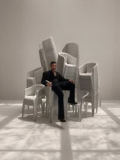 a man sitting in a chair surrounded by stacks of white plastic chairs, with his legs crossed