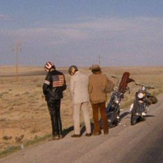 three men standing on the side of a road next to a motorcycle and two motorcycles