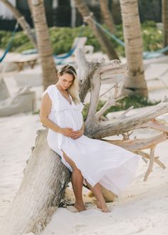 a pregnant woman sitting on top of a wooden log in the sand next to palm trees