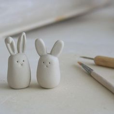 two white ceramic rabbits sitting next to each other on a table with paintbrushes