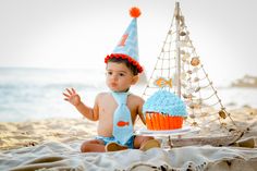 a little boy sitting on the beach with a cupcake in front of him wearing a birthday hat