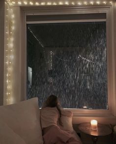 a woman sitting on a couch in front of a window covered in rain falling from the sky