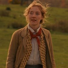 a woman standing in front of a field with grass and trees behind her, wearing a brown jacket and red bow tie