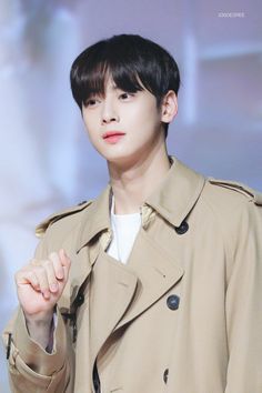 a young man in a trench coat holding his hand up to the side while standing next to a wall