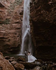 a bride and groom standing in front of a waterfall at the base of a cliff