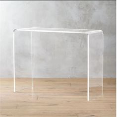 an acrylic table with clear legs on a wooden floor in front of a concrete wall