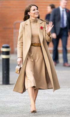 a woman in a tan coat is walking down the street with her hand up and smiling