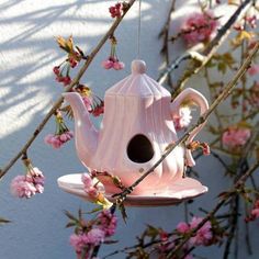 a pink teapot hanging from a tree branch with flowers in the foreground and a white wall behind it