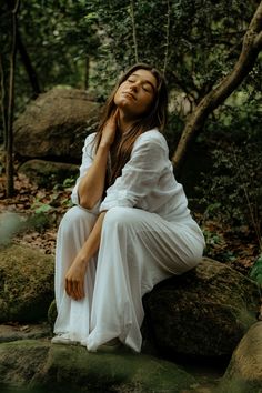 a woman sitting on top of a rock in the forest with her eyes closed and hands under her chin