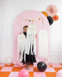 a woman standing in front of a pink wall holding up two white ghost decorations with balloons and streamers